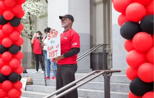 A CFA member holds a sign that reads “stop the rain of debt” at a rally with red and black balloons.