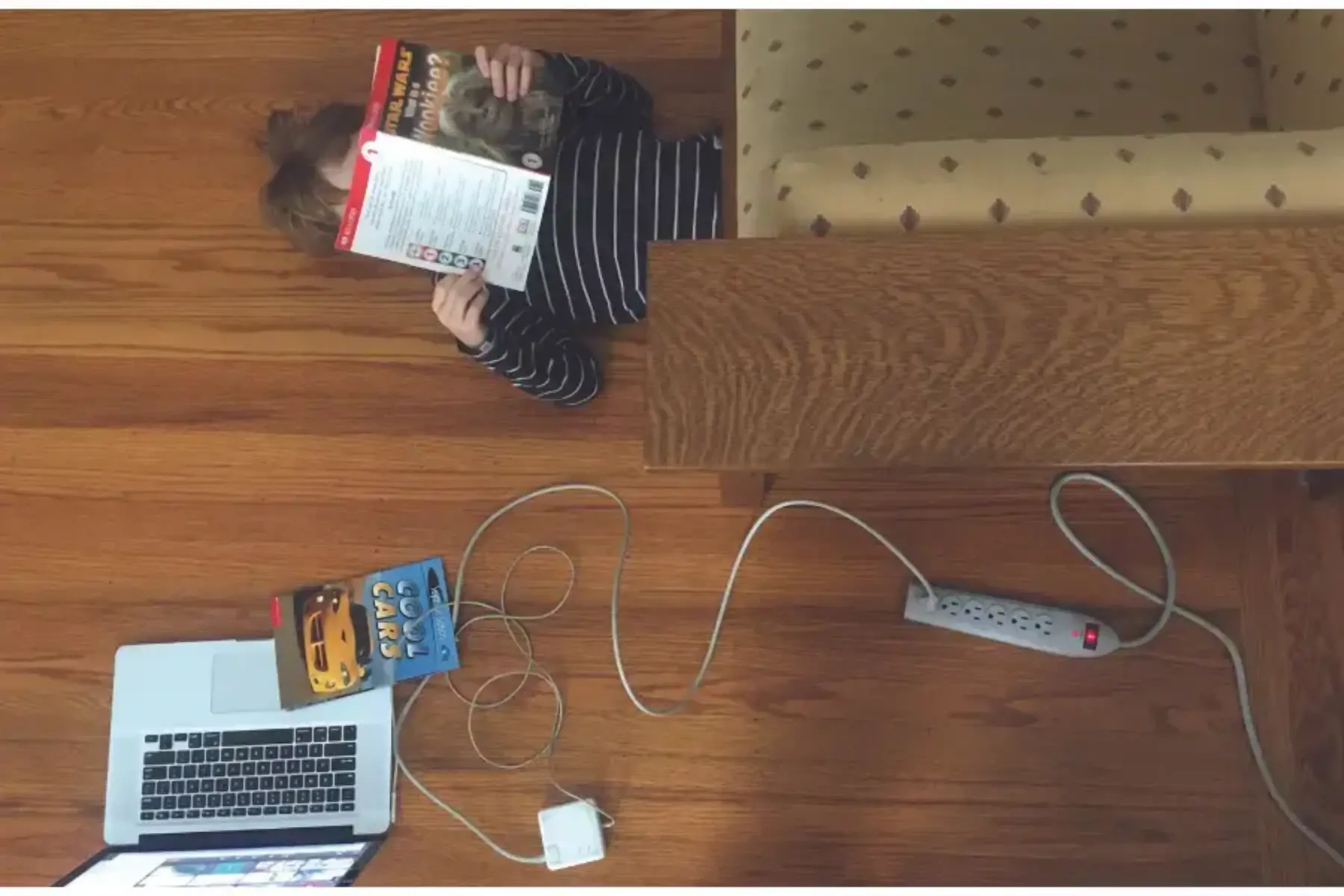 Photo shot from ceiling looking down on a child laying down on the ground with their legs under the couch. The child has brown hair, is wearing a black and white striped shirt, and is reading a Star Wars book. The floor is a dark wood floor. The couch is dark wood with olive green fabric. Next to the child on the floor is a power strip, an open laptop with a Zoom meeting. The laptop is charging via the power strip. And a Cool Cars book lies on top of the laptop keyboard.