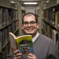 A headshot of a person wearing glasses, a grey sweater over a white collared button-up, standing in between library bookshelves while holding a book.