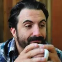 An off-guard photo of a person holding a white coffee mug looking to the right side. They are wearing a blue plaid button-up.