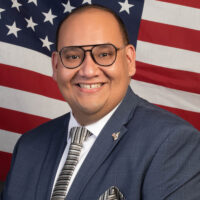Candidate with blue suit jacket, tie, and American flag background.