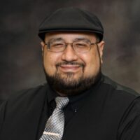 man wearing a hat, tie, and glasses smiles in front of a black backdrop