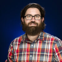 A man with dark brown hair and black glasses in a red, blue and orange plaid T-shirt smiles for a photo with a dark blue background