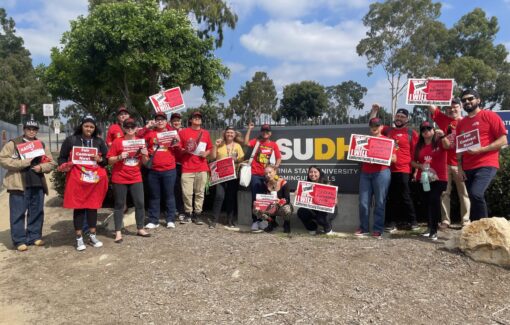 Faculty and students in red CFA shirts in front of the CSUDH sign hold signs in preparation for 2024's Strike.