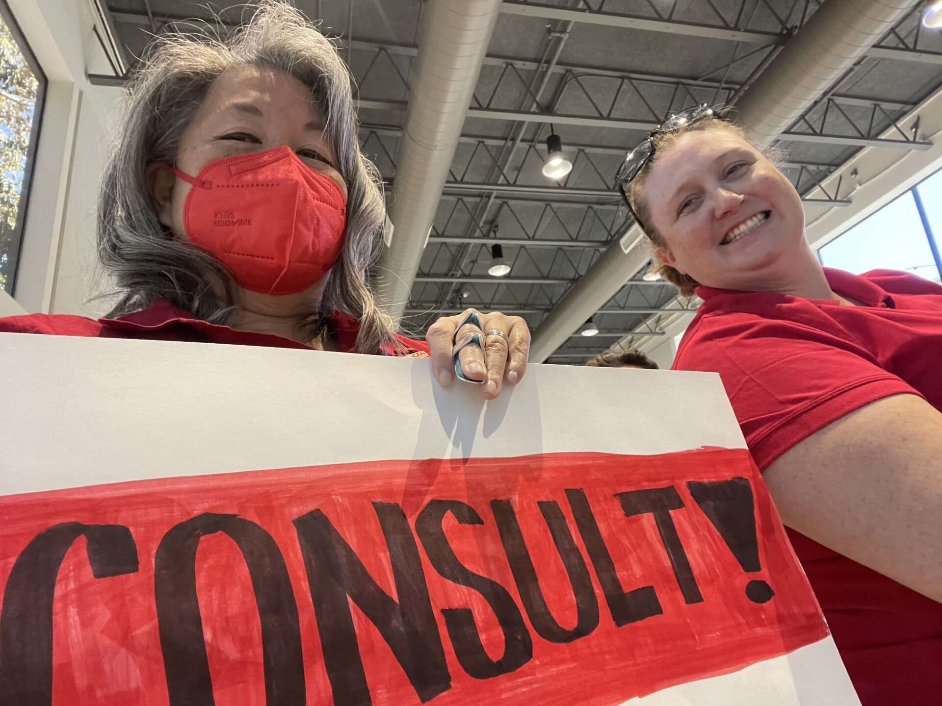 two women in photo, one holding a sign reading "consult" in black text on red background.