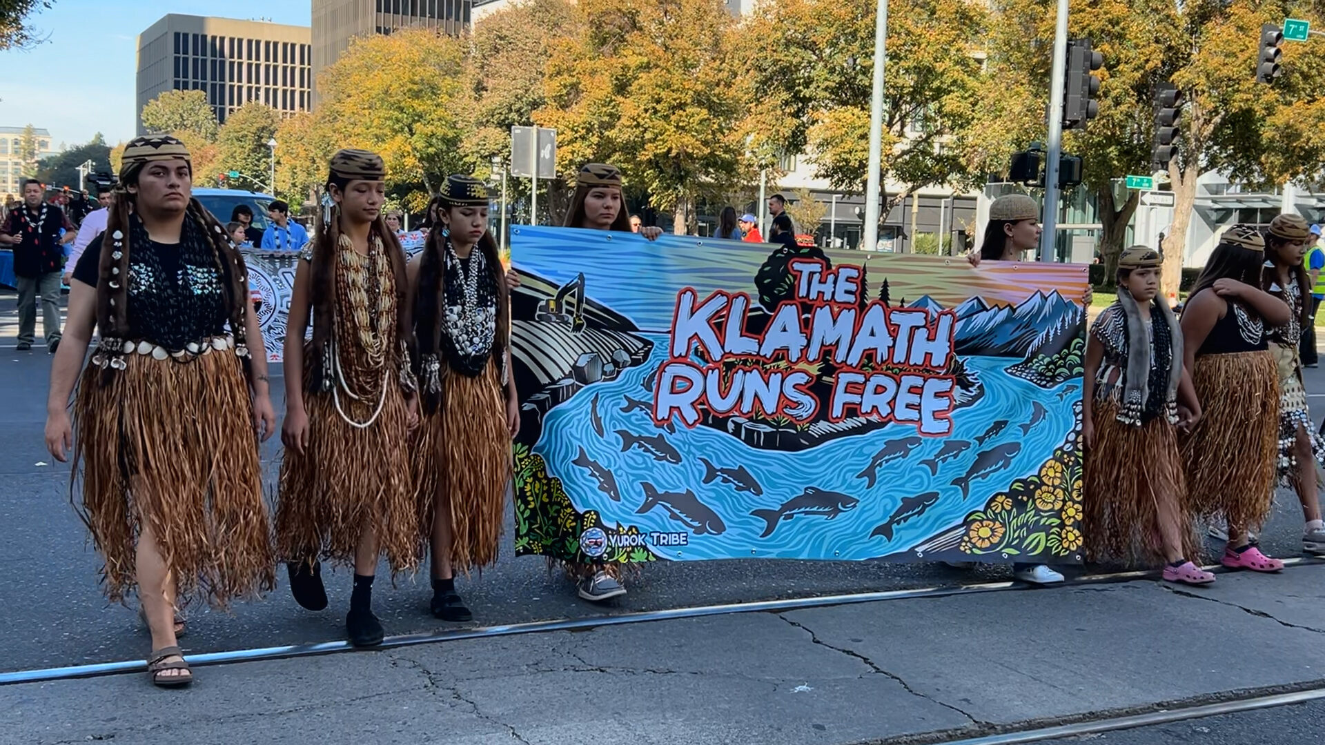 Native people wear native attire and hold a banner saying The Klamath Runs Free.