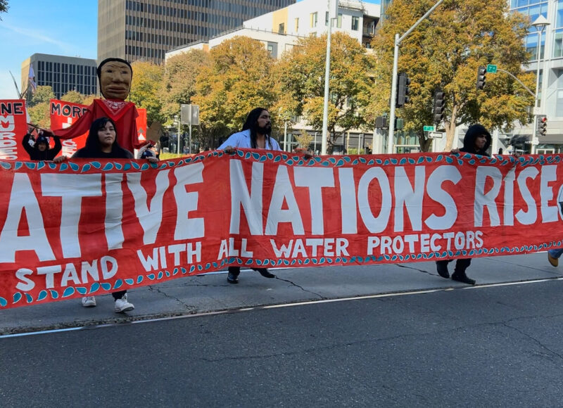 Native people carry a red banner with white text that reads Native Nations Rise. Stand with all Water Protectors.