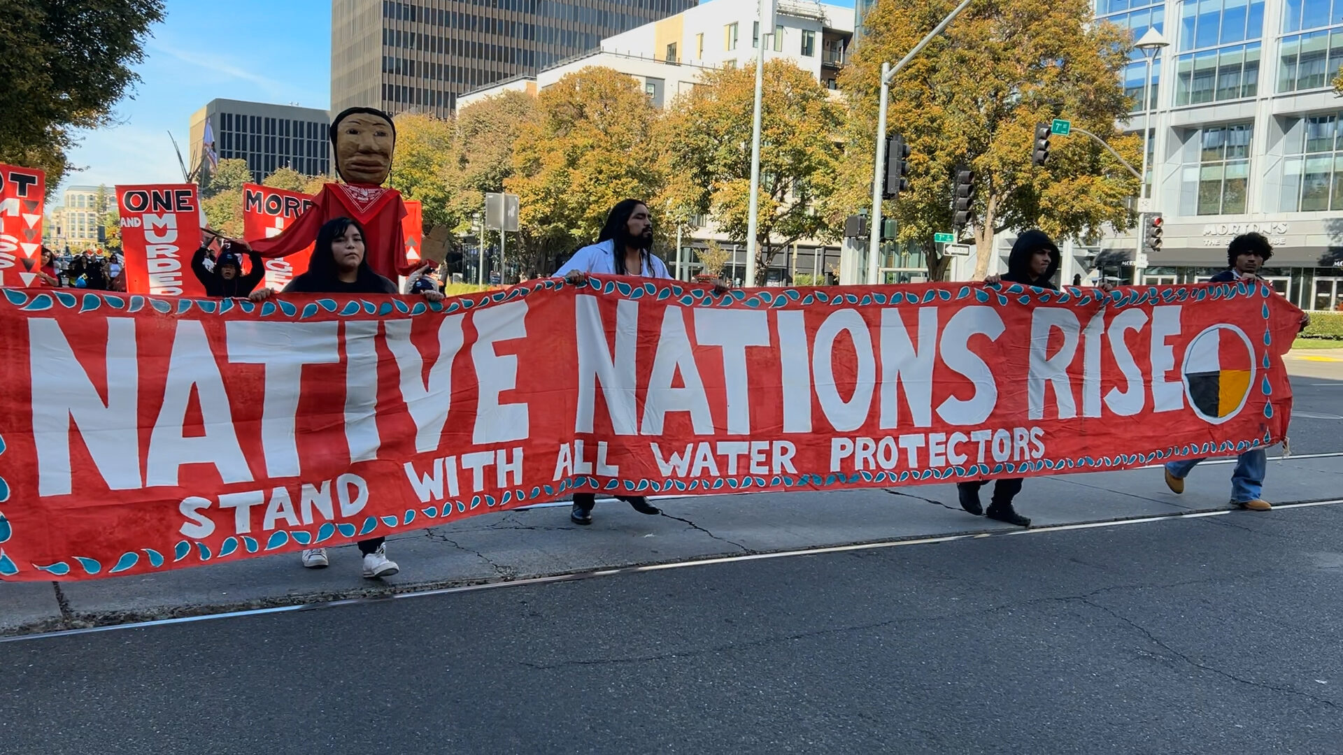 Native people carry a red banner with white text that reads Native Nations Rise. Stand with all Water Protectors.