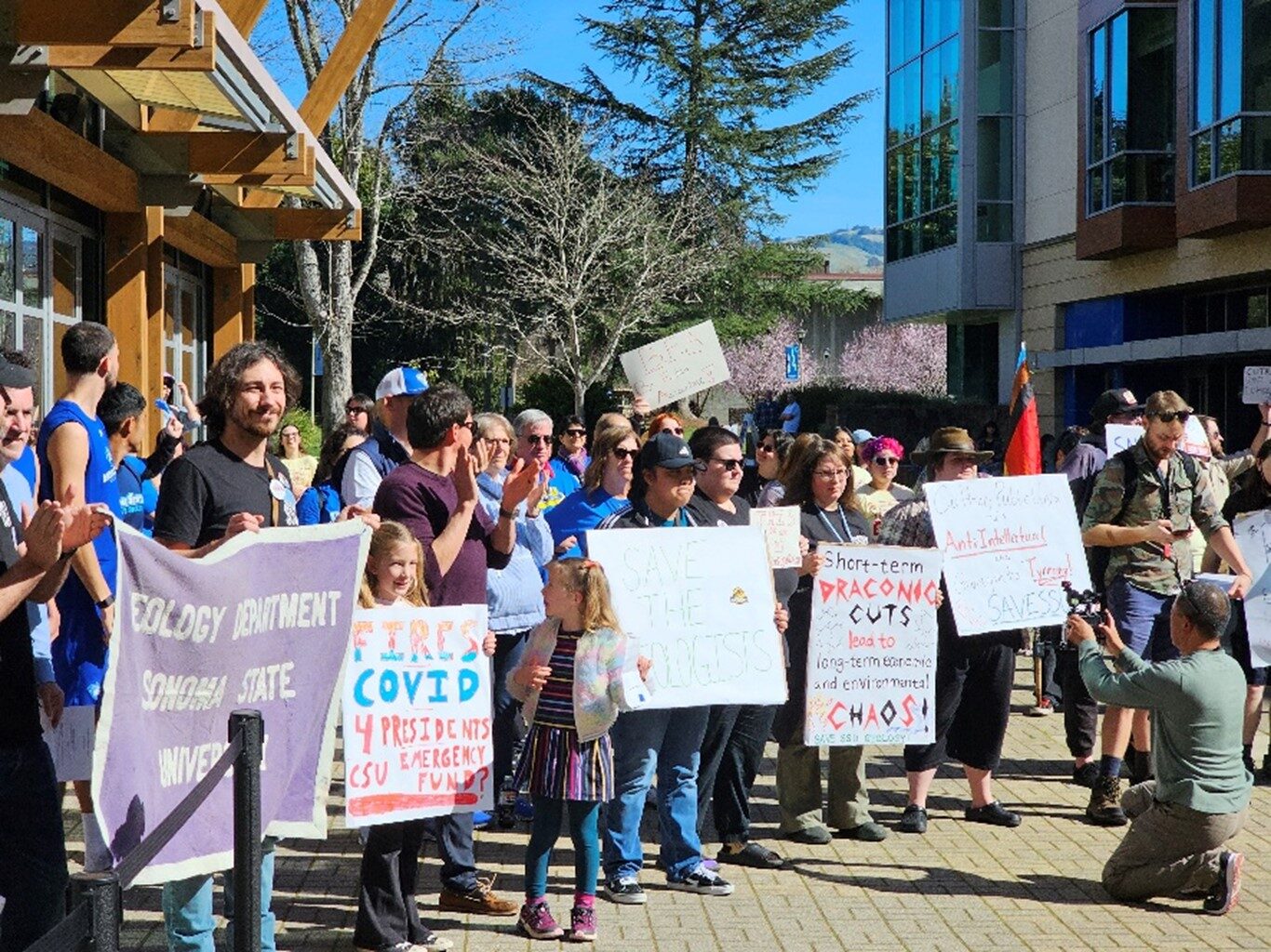 A group of people outdoors with signs and banners demanding sonoma state management to stop the layoffs