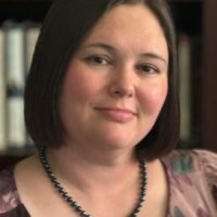 Headshot of Library Department Representative for the Union: Rachel Keiko Stark. She is pictured in front of a bookcase, wearing a patterned mauve shirt, dark pearl, single-strand necklace, and has a brunette bob.